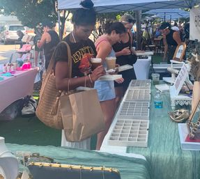 Women browsing jewelry trays at an outdoor artisan weekend market, holding iced coffees and shopping bags under vendor canopies