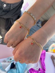 Close-up of two wrists wearing matching gold paperclip bracelets with small red cherry charms, stacked with a geometric gold-and-silver bracelet and rings over denim and colorful swimwear — summer women's jewelry accessory