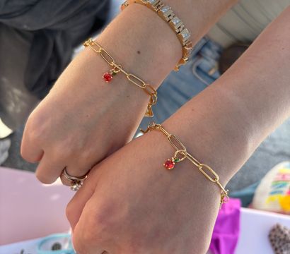 Close-up of two wrists wearing matching gold paperclip bracelets with small red cherry charms, stacked with a geometric gold-and-silver bracelet and rings over denim and colorful swimwear — summer women's jewelry accessory