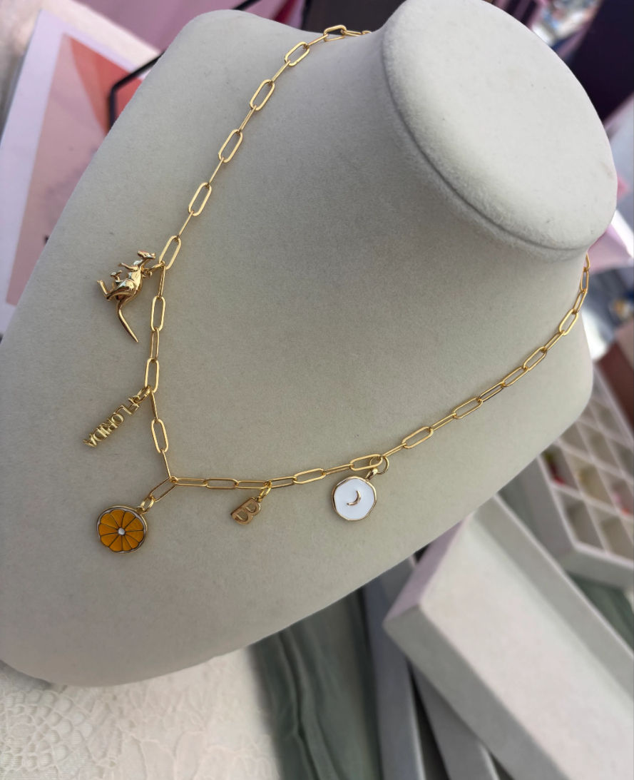 Gold paperclip-chain charm necklace on a beige display bust featuring an orange enamel citrus slice, white enamel crescent moon, small gold initial and tag — dainty boutique jewelry display.
