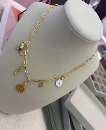 Gold paperclip-chain charm necklace on a beige display bust featuring an orange enamel citrus slice, white enamel crescent moon, small gold initial and tag — dainty boutique jewelry display.