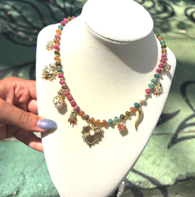 Multicolored beaded charm necklace on a white display bust, gold-tone charms including a filigree heart with pearl, sun, horn and dangling beads — vibrant boho jewelry