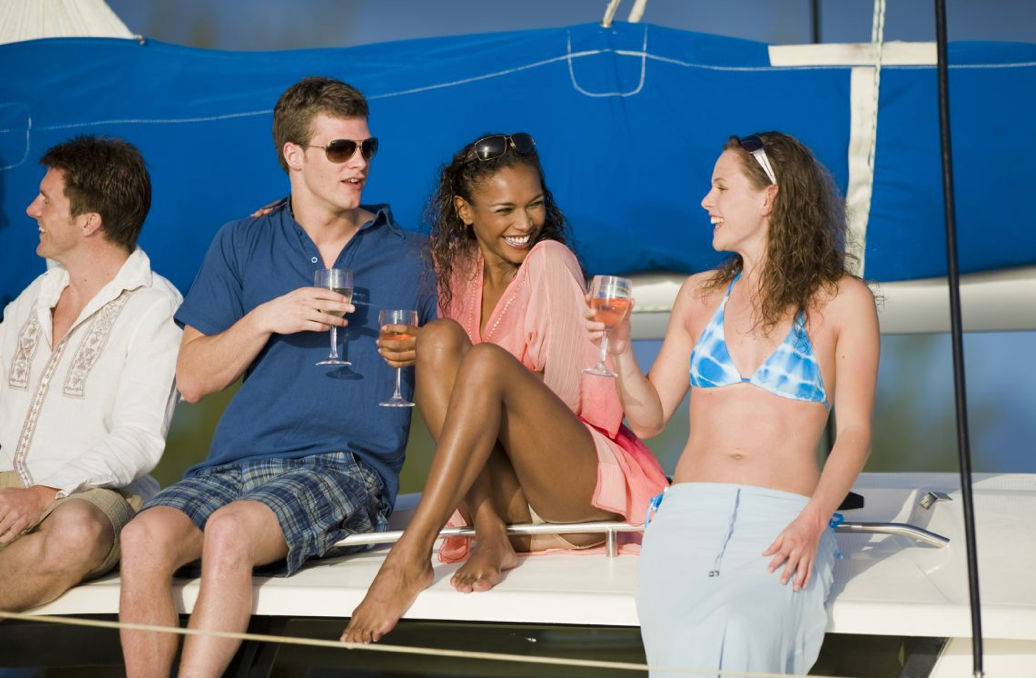 Four friends laughing on a sailboat deck in swimsuits and summer clothes, toasting with wine glasses under a bright blue sail on a sunny coastal getaway