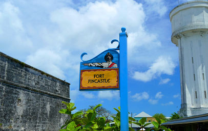 Colorful blue post sign reading 'Fort Fincastle' with a painted pointing soldier, set beside an old stone fort wall and a white tower under a bright blue sky.
