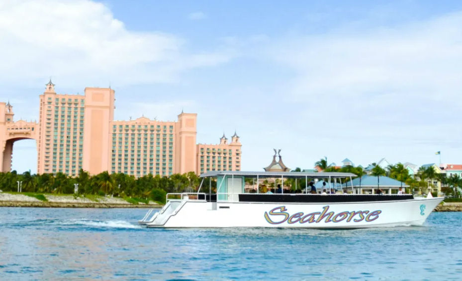 White sightseeing boat with colorful lettering cruising on blue tropical water past a palm-lined shore and a large coral-pink beachfront resort with towers and arch under a sunny sky