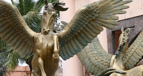 Bronze winged-horse fountain (Pegasus) with outstretched wings and water spray, green patina and palm tree against a peach stucco building