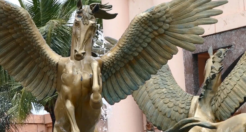 Bronze winged-horse fountain (Pegasus) with outstretched wings and water spray, green patina and palm tree against a peach stucco building