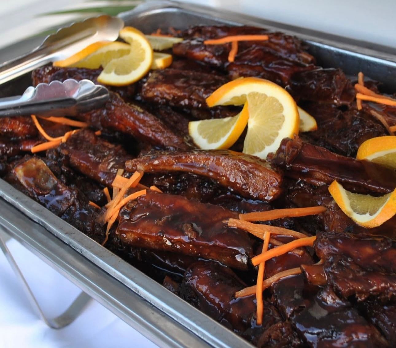 Sticky glazed barbecue ribs piled in a stainless-steel catering tray, garnished with orange slices and shredded carrots with metal serving tongs