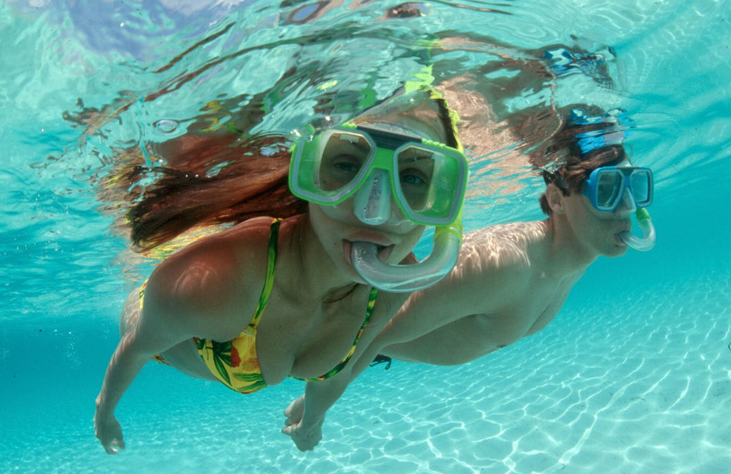 Two snorkelers wearing bright masks glide underwater in clear turquoise water above a sunlit rippled sandy seabed, woman in a floral bikini in front and a man close behind.