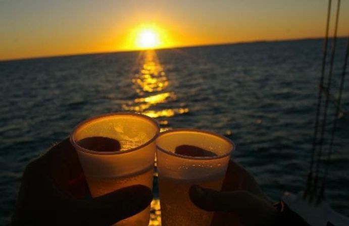 Two plastic cups of beer clinking on a sailboat at sunset with the golden sun reflecting across the ocean