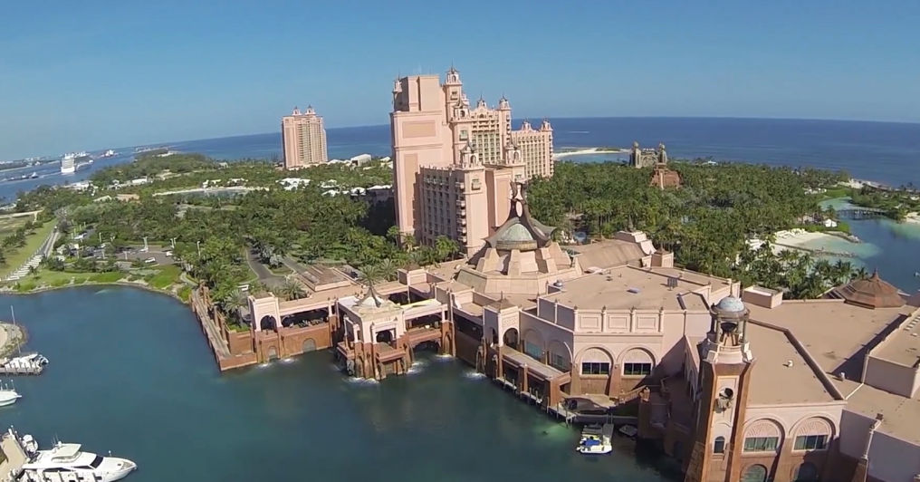Aerial view of a pink-hued luxury island resort with tall hotel towers, marina with yachts, turquoise lagoons, dense palm trees and Atlantic Ocean horizon.