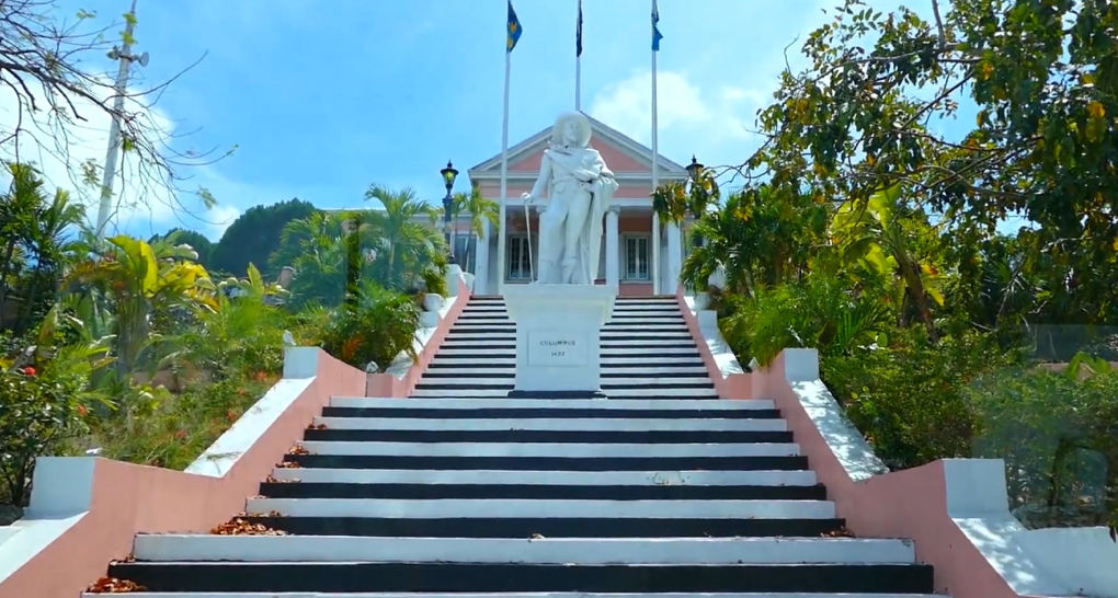 Sunlit tropical staircase with black-and-white steps leading up through palm trees to a pink neoclassical building topped by a white robed statue and flagpoles against a bright blue sky.