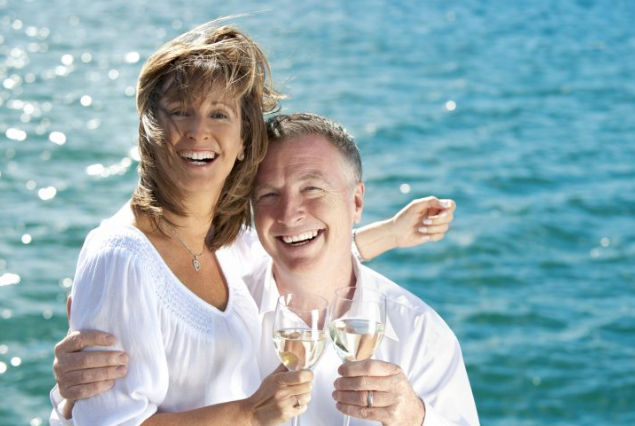 Smiling couple in white shirts toasting with white wine glasses by the sunlit blue ocean, wind-swept hair and joyful expressions