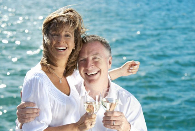 Smiling couple in white shirts toasting with white wine glasses by the sunlit blue ocean, wind-swept hair and joyful expressions