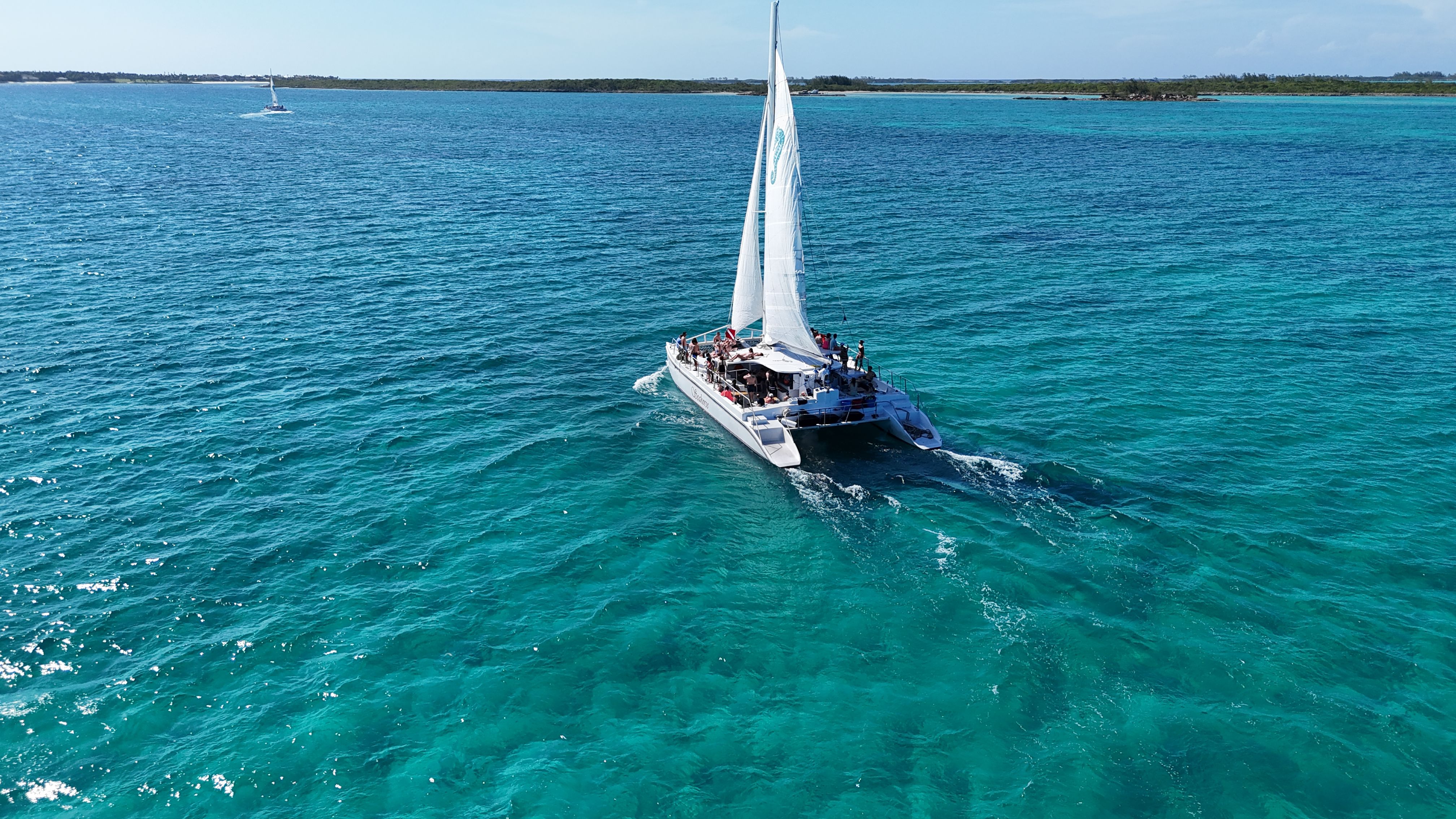 Sunlit white catamaran with sails up carrying passengers across crystal-clear turquoise tropical waters toward a low-lying island under a blue sky.