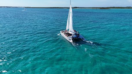 Sunlit white catamaran with sails up carrying passengers across crystal-clear turquoise tropical waters toward a low-lying island under a blue sky.