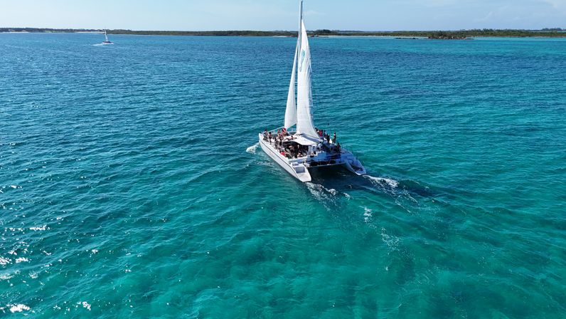 Sunlit white catamaran with sails up carrying passengers across crystal-clear turquoise tropical waters toward a low-lying island under a blue sky.