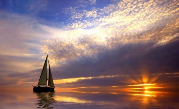 Sailboat on calm ocean at golden sunset with dramatic cloud-filled sky and sun rays reflecting on glassy water.