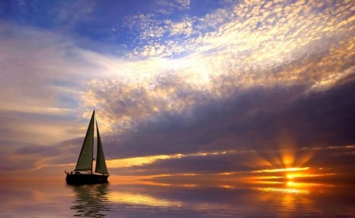 Sailboat on calm ocean at golden sunset with dramatic cloud-filled sky and sun rays reflecting on glassy water.