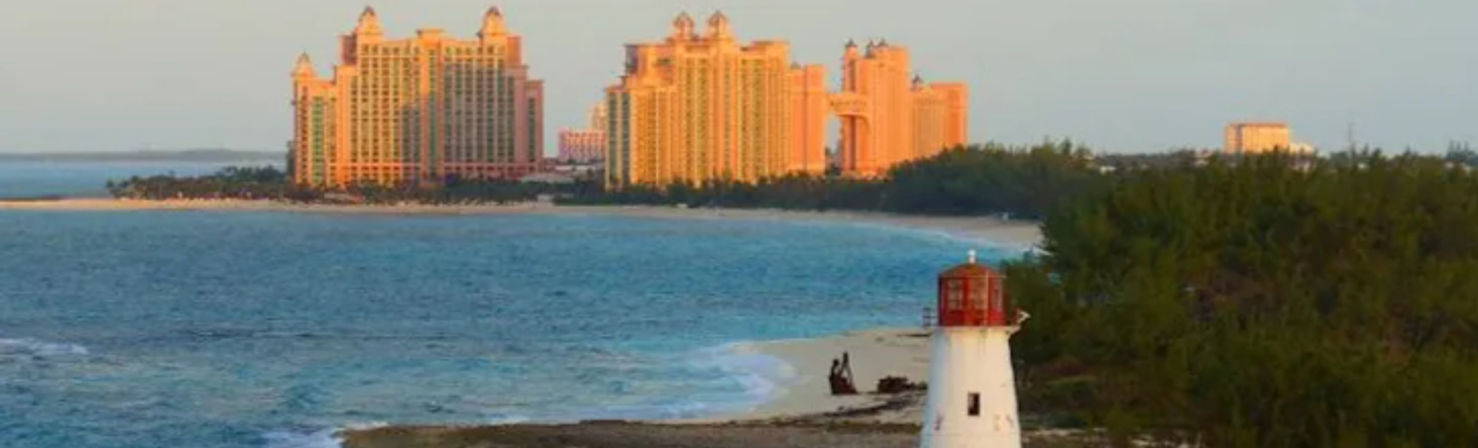 Sunset view of a white lighthouse on a tropical sandy shore with turquoise water and lush greenery, luxury beachfront resort towers glowing orange on the distant skyline