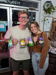 Smiling young couple holding hand-painted beer mugs featuring colorful gnome designs inside a cozy craft brewery or taproom.