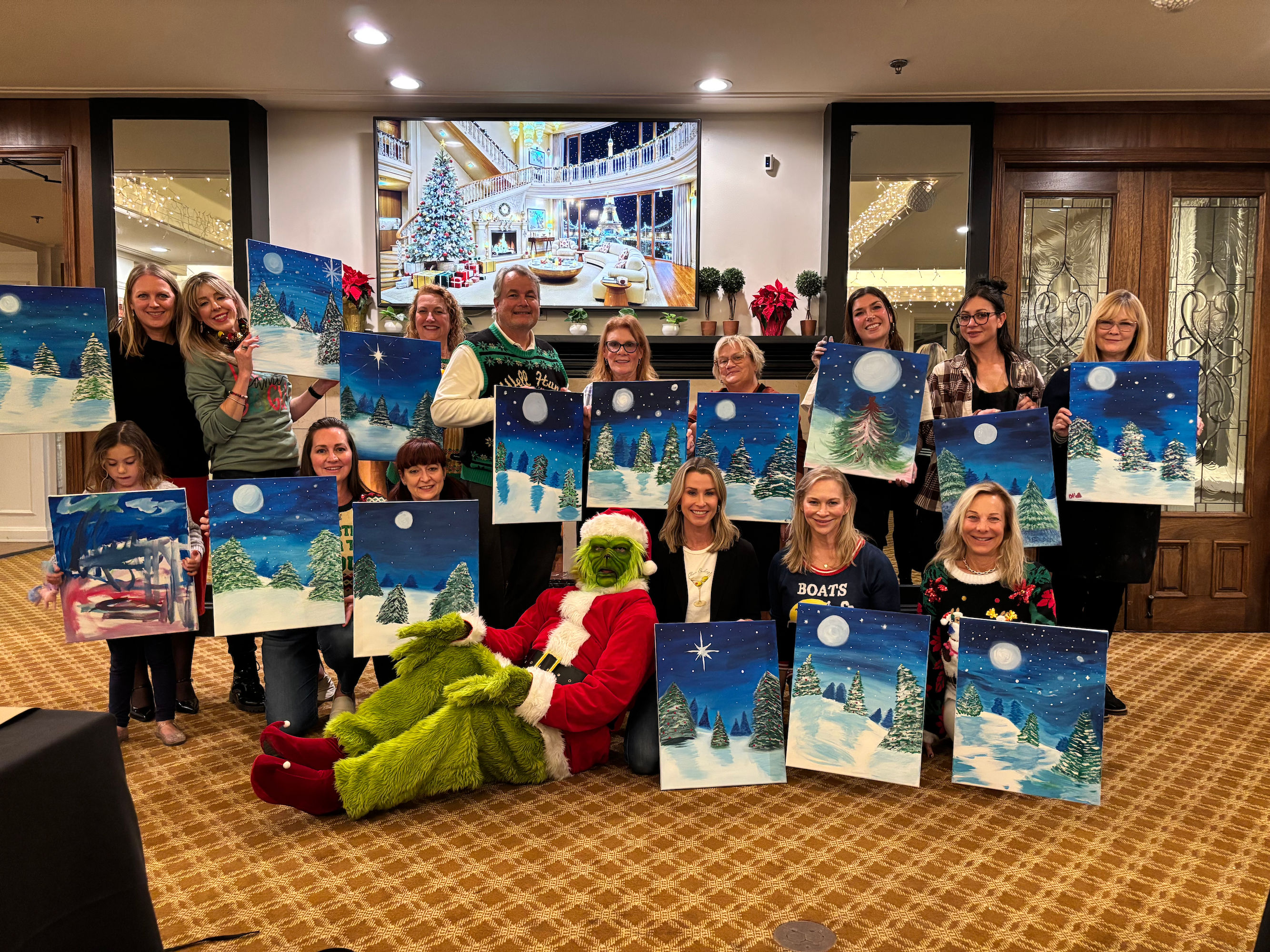 Group holiday paint night in a hotel lobby, people and a child holding moonlit snowy-tree canvas paintings, with someone dressed as the Grinch in a Santa suit seated in front and festive decorations in the background.