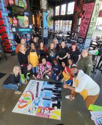 Diverse group of people smiling and kneeling around a large colorful jigsaw puzzle that reads “Indianapolis” on the floor of a mural-covered restaurant or brewery with high windows and tables, a fun team-building community event.