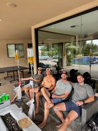 Four friends relaxing on a covered patio couch by a backyard pool, IV hydration bags on poles, ping-pong table and glass doors in the background