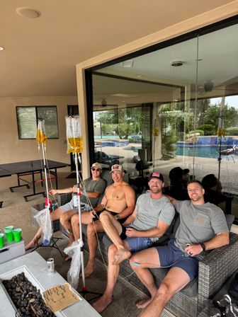 Four friends relaxing on a covered patio couch by a backyard pool, IV hydration bags on poles, ping-pong table and glass doors in the background