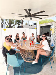 Group of eight women smiling around a wicker patio table under a large ceiling fan on a sunny backyard covered patio, sipping drinks while IV hydration bags hang from stands.
