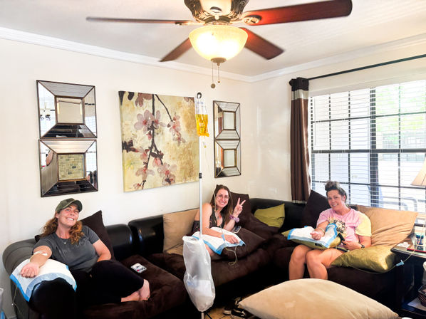 Three people relaxing on a home living-room sectional receiving IV infusions from a yellow bag on a pole, with floral wall art, decorative mirrors, a ceiling fan and a sunlit window.