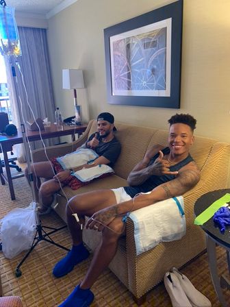 Two smiling young men relaxing on a striped hotel-room couch receiving IV drip hydration, arms connected to IV lines with an IV pole, side table, lamp and framed artwork in the background.