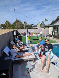 Friends relaxing with IV hydration drips poolside at a sunny Southwest suburban backyard party with lounge chairs, balloons, palm trees and a swimming pool
