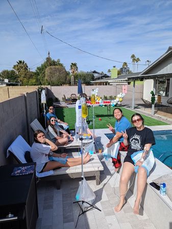 Friends relaxing with IV hydration drips poolside at a sunny Southwest suburban backyard party with lounge chairs, balloons, palm trees and a swimming pool