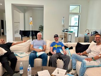 Four men sitting and smiling on a beige sectional in a bright modern living room, each hooked up to IV hydration bags on poles, with water bottles, coffee cup and paperwork on a coffee table in the foreground.