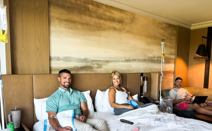 Three adults relaxing in a modern hotel room receiving IV drip therapy — two propped up on a bed smiling and one lounging on a sofa with a laptop, bedside IV poles and cozy decor visible.