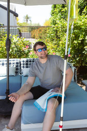 Smiling man in sunglasses relaxing on a blue patio sofa under an umbrella in a sunny backyard garden while receiving an IV infusion from a hanging bag.