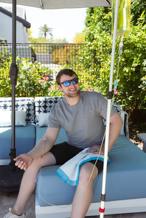 Smiling man in sunglasses relaxing on a blue patio sofa under an umbrella in a sunny backyard garden while receiving an IV infusion from a hanging bag.