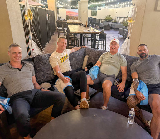 Four men lounging on an outdoor patio couch under string lights, each hooked up to IV drip bags during a casual wellness evening.