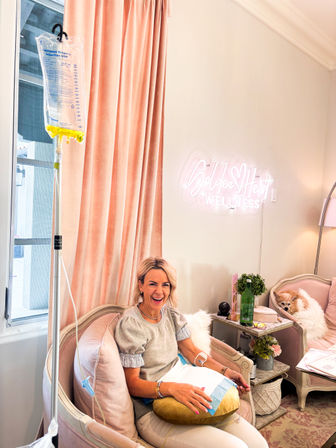 Smiling woman getting IV therapy in a pink, spa-like wellness lounge with velvet curtains, plush chairs, a side table with plants and a small dog napping on the adjacent seat.