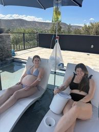 Two women in swimsuits reclining on a rooftop pool deck under an umbrella receiving IV hydration drips, with scenic arid hills in the background.