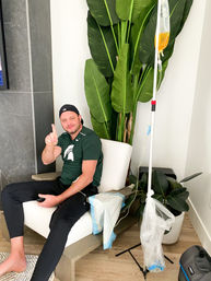 Smiling man in a cap lounging in a modern home living room chair receiving IV therapy from a yellow infusion bag on a pole, pointing up next to a large potted tropical plant.