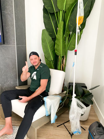 Smiling man in a cap lounging in a modern home living room chair receiving IV therapy from a yellow infusion bag on a pole, pointing up next to a large potted tropical plant.