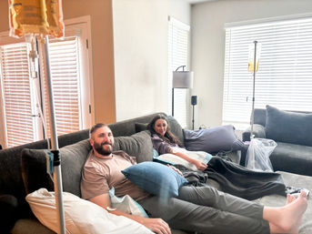 Smiling man and woman lounging on a gray sectional in a sunlit living room, each receiving at-home IV hydration drips from poles beside the couch with pillows and blankets.