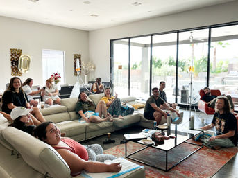 Group of people lounging on a large sectional in a sunlit modern living room with floor-to-ceiling sliding glass doors, chatting and smiling while connected to IV stands and drip bags during a relaxed communal treatment session.