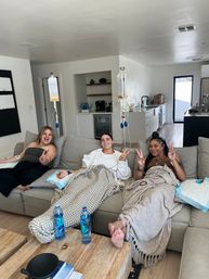 Three friends lounging on a living room sofa receiving IV hydration drips, wrapped in blankets and flashing peace signs, with water bottles and IV bags nearby — at-home wellness session.