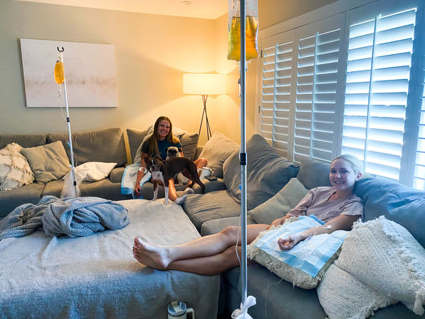 Two people relaxing on a grey sectional in a cozy living room, each receiving IV fluids from poles, accompanied by two small dogs, soft blankets, a floor lamp and white plantation shutters.