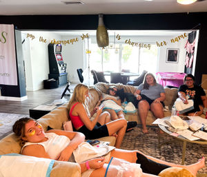 Bachelorette spa party at home: friends lounging on a living-room sectional with under-eye patches and leg masks, a gold champagne balloon and banner reading pop the champagne she's changing her name overhead.