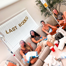 Four women lounging in a cozy living room under a 'LAST RODEO' banner, smiling while receiving IV hydration drips from IV bags on poles, with blankets, cups and party supplies on the coffee table.