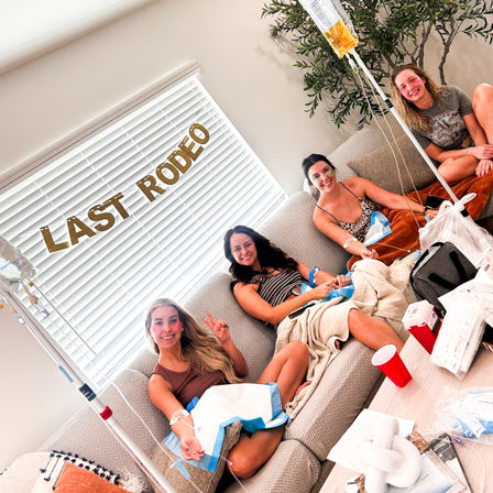Four women lounging in a cozy living room under a 'LAST RODEO' banner, smiling while receiving IV hydration drips from IV bags on poles, with blankets, cups and party supplies on the coffee table.